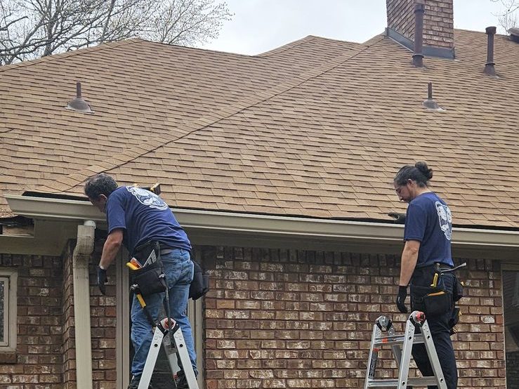 Two installers on ladders at the roofline of a brick home